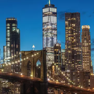 The photo captures the  Brooklyn Bridge at night from Brooklyn, with illuminated city lights reflecting on the calm water. The skyline, a modern skyscraper, and the serene park enhance the scene., License Type: media, Download Time: 2025-11-26T20:58:49.000Z, User: LP_YKhanna, Editorial: false, purchase_order: 65050 - Digital Destinations and Articles, job: LP, client: App Content, other: Yuvraj Khanna