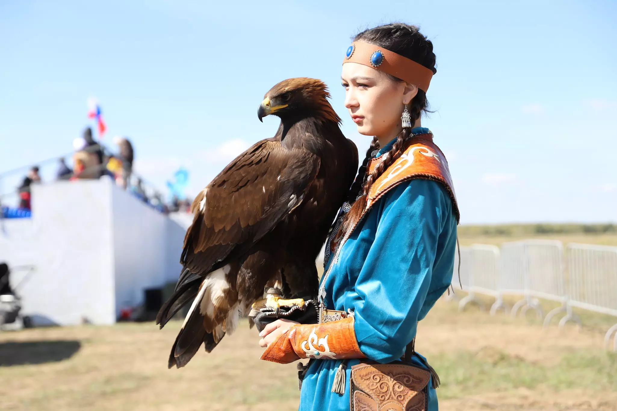 A woman in a traditional blue tunic and leather headband holds a falcon during a competition.