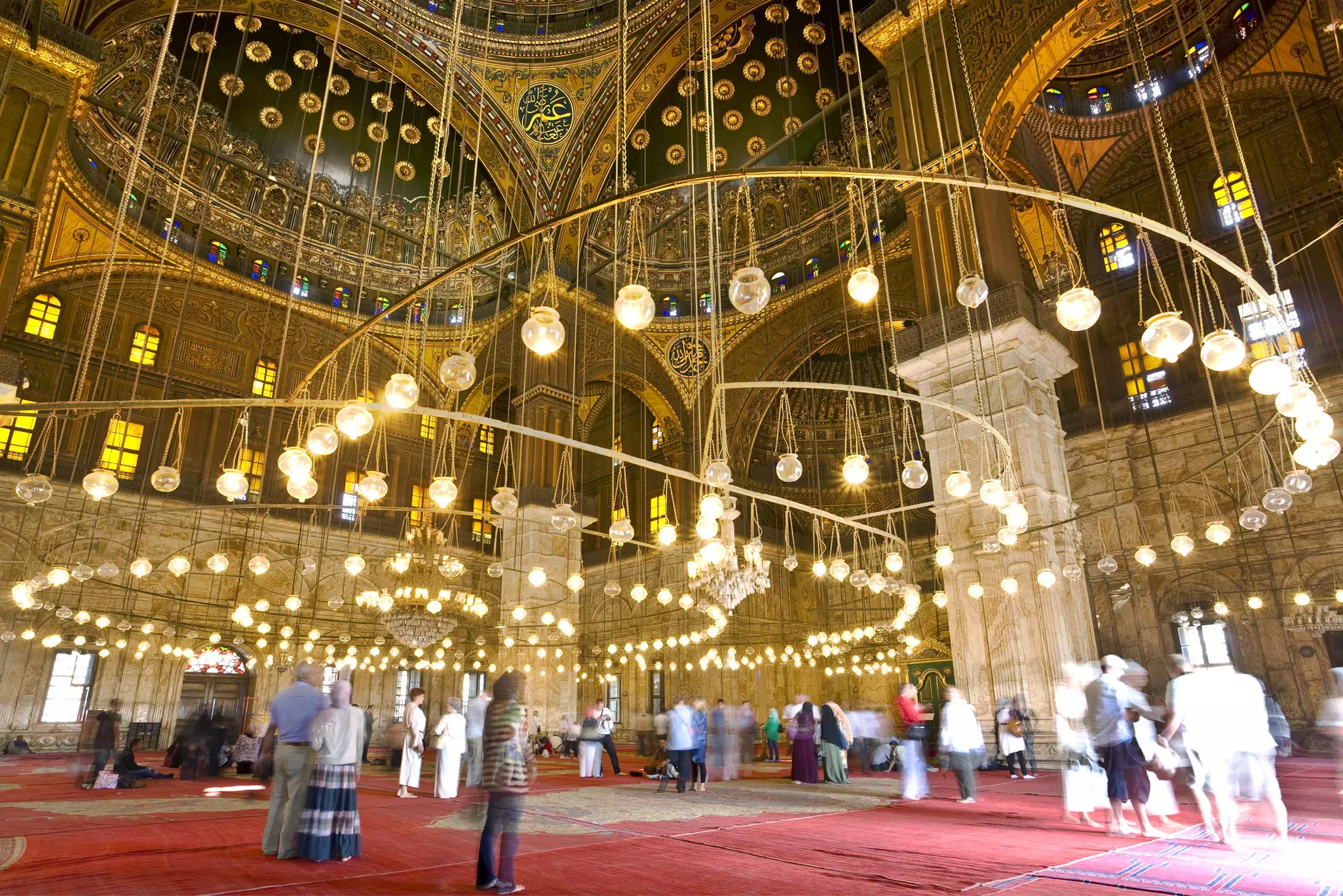 People admiring the chandeliers and ceilings of a vast mosque.
