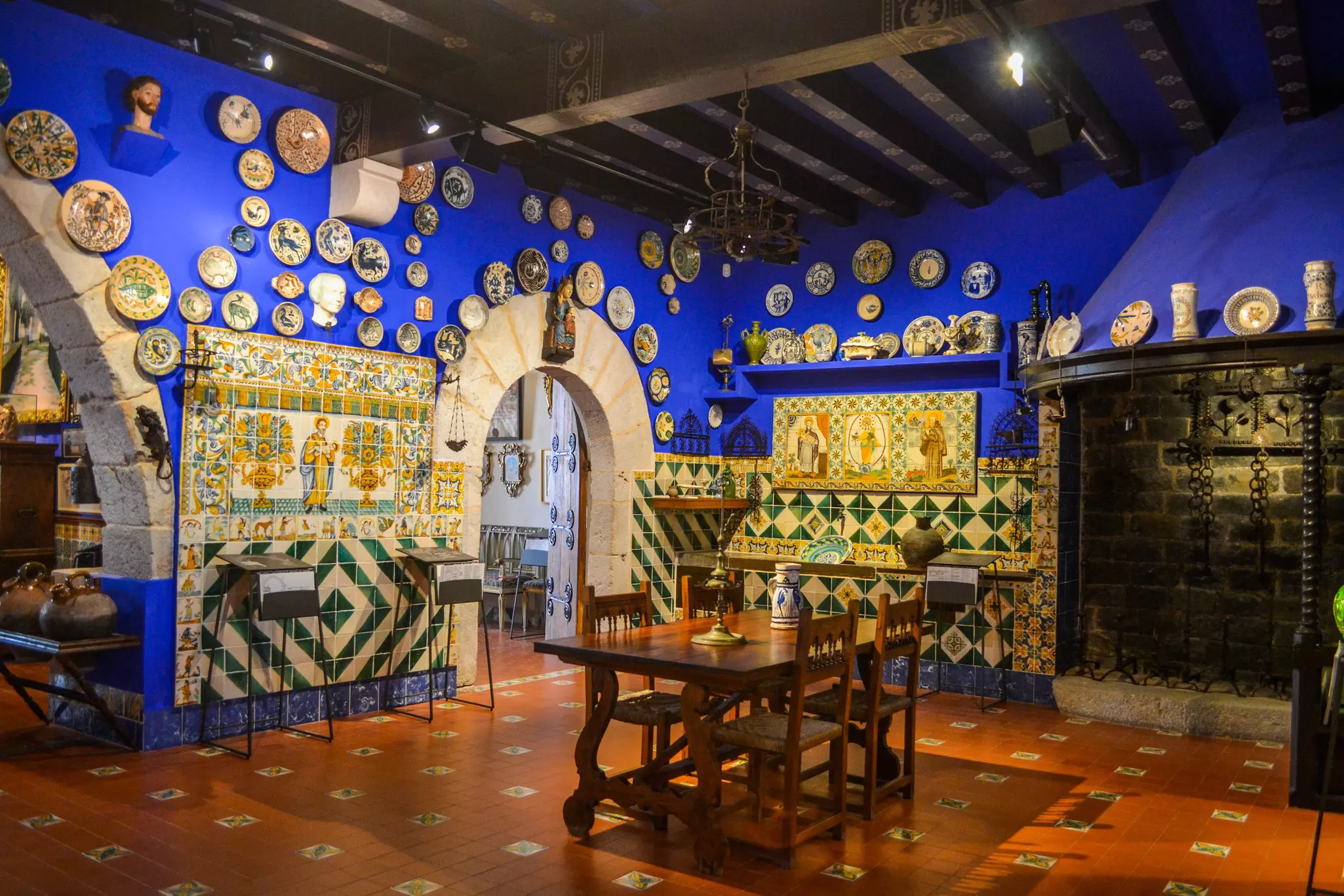 A room with vivid blue walls lined with ornate plates and detailed tiles of people and plants. A wooden table with four chairs is in the middle of the room, whose floor has a orange hue.