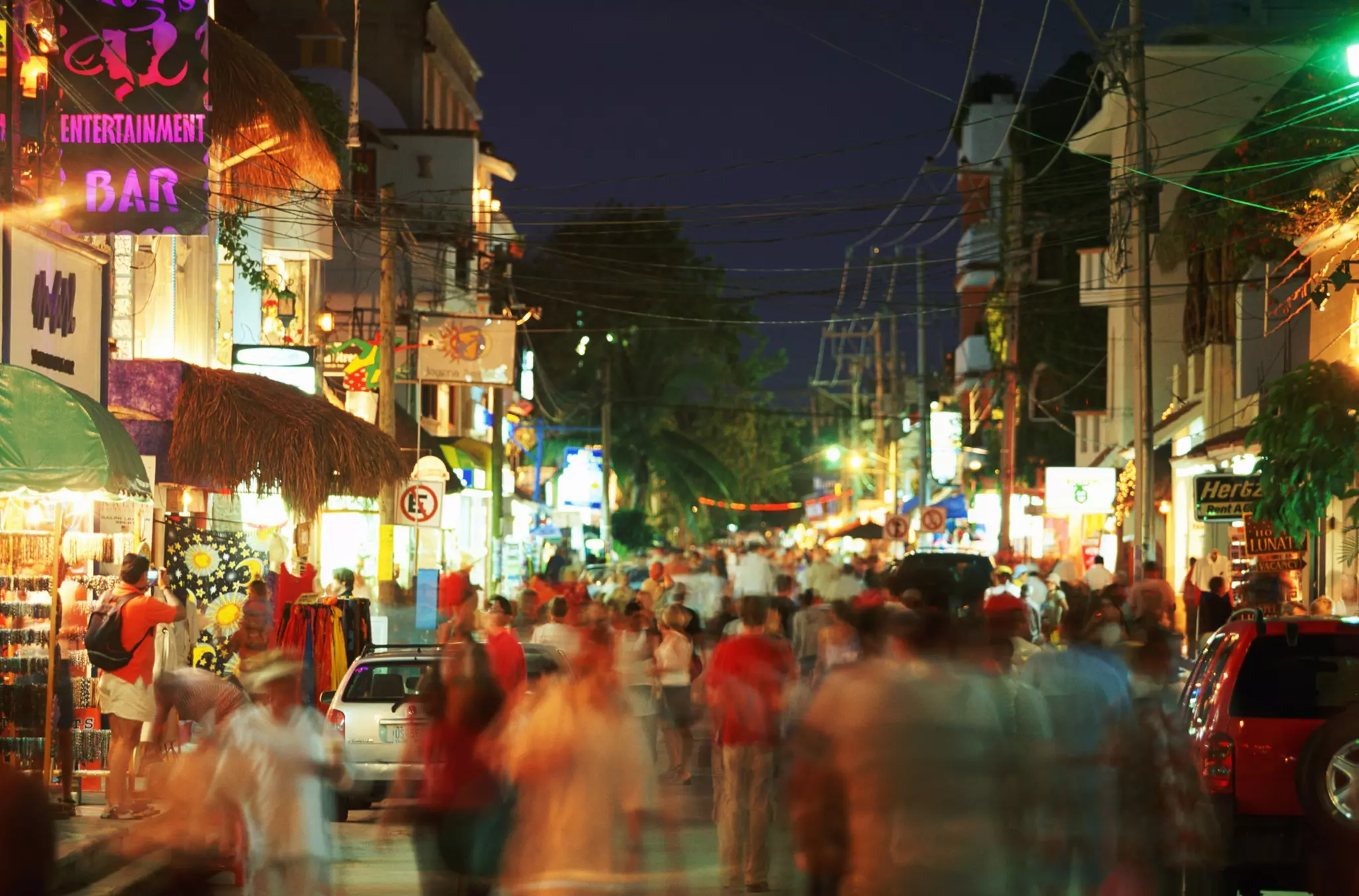 People appear blurry as they walk down a city street at night. Signs of restaurants and shops illuminate the street.