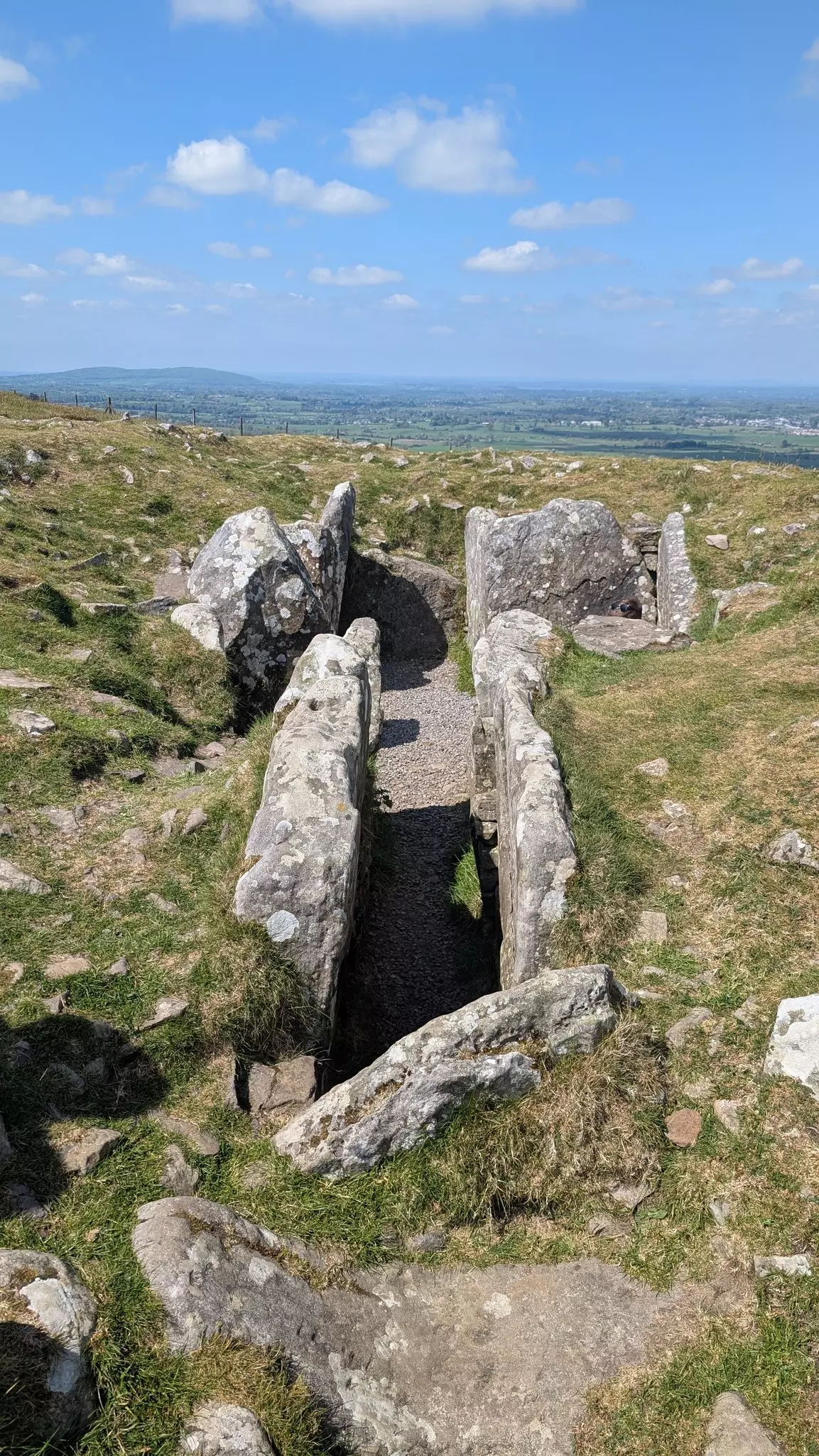 Large slabs of stone mark the entrance to an ancient hilltop tomb, with views of the countryside rolling into the distance.