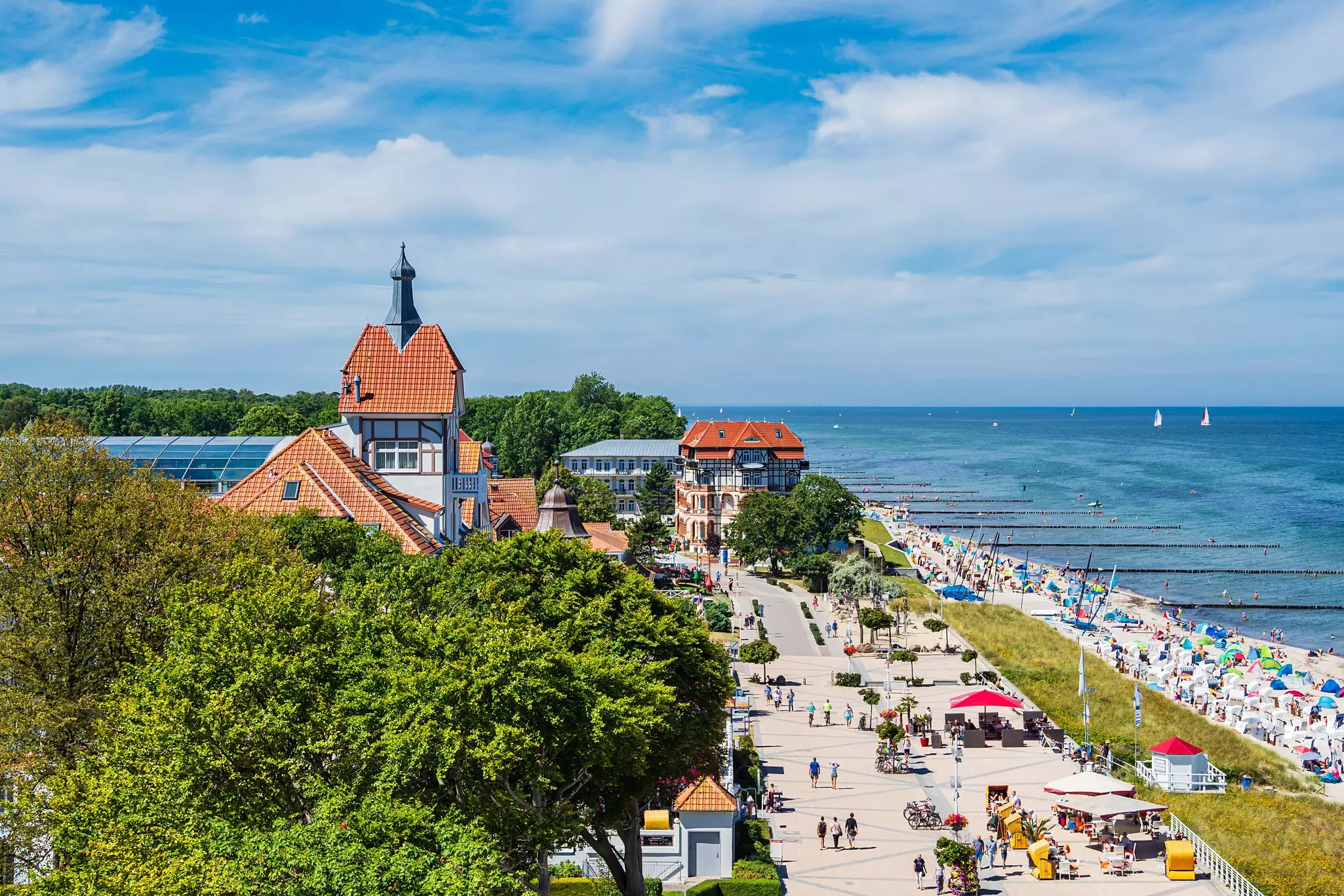A busy seafront in a resort town, with stalls and rental options lining the beach as people walk along the promenade.