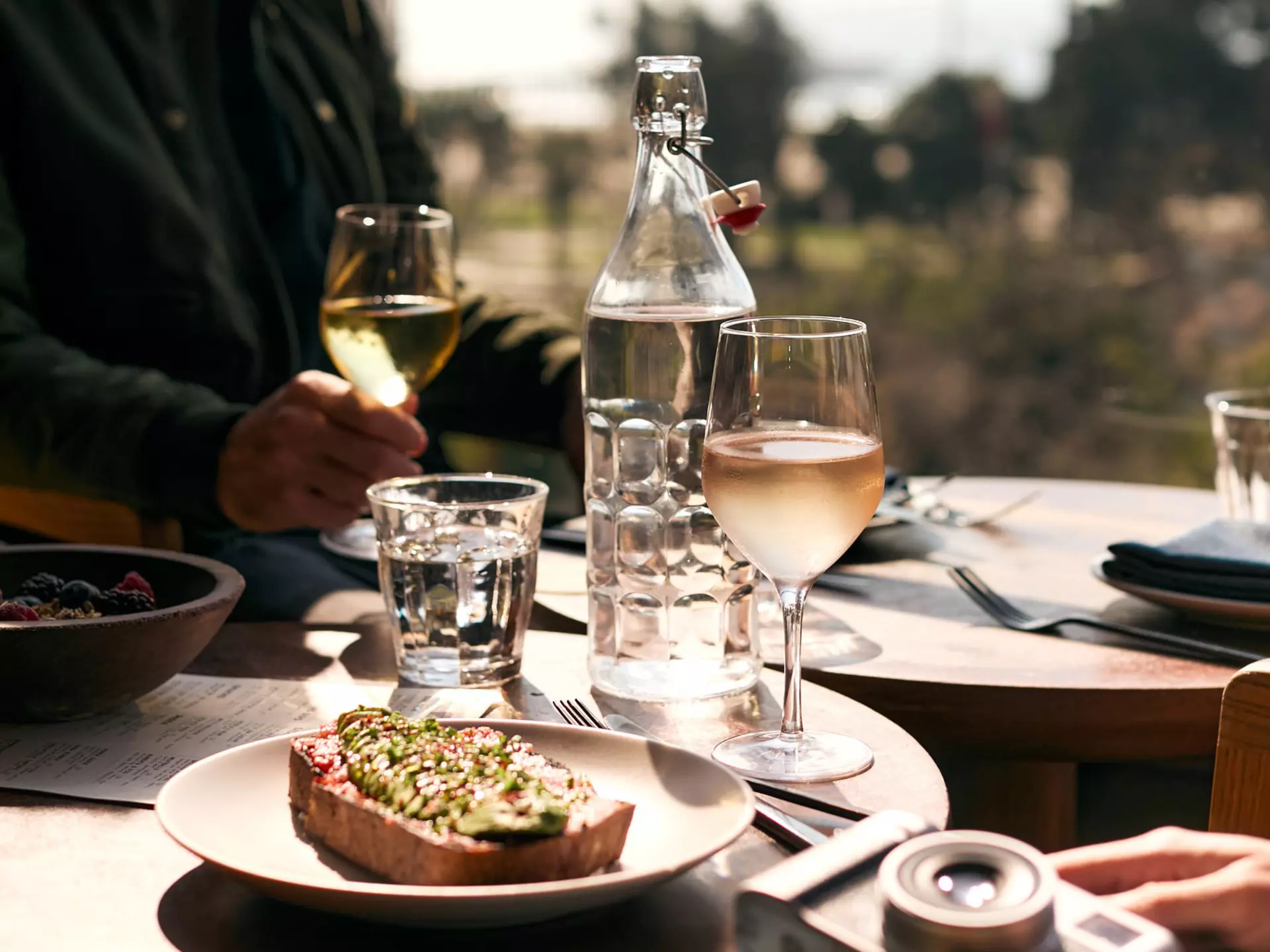 A close shot of a dish on a table with wine glasses and water nearby. In the upper right, a man is looking out at the ocean view, and a woman's hand with a camera is visible in the bottom right corner
