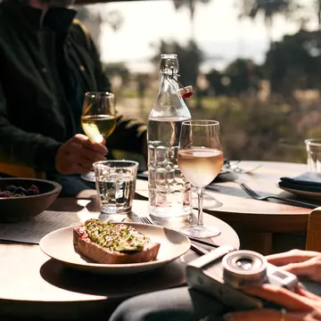 A close shot of a dish on a table with wine glasses and water nearby. In the upper right, a man is looking out at the ocean view, and a woman's hand with a camera is visible in the bottom right corner