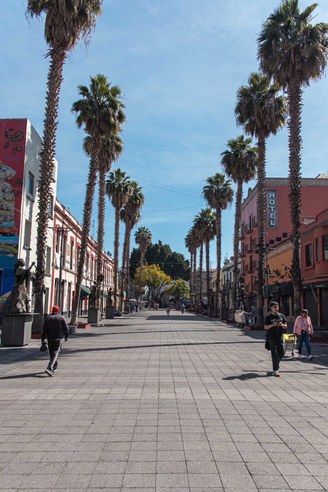 View down a pedestrian street by Plaza Garibaldi, lined with tall palm trees and colorful buildings under a bright blue sky in Mexico City.