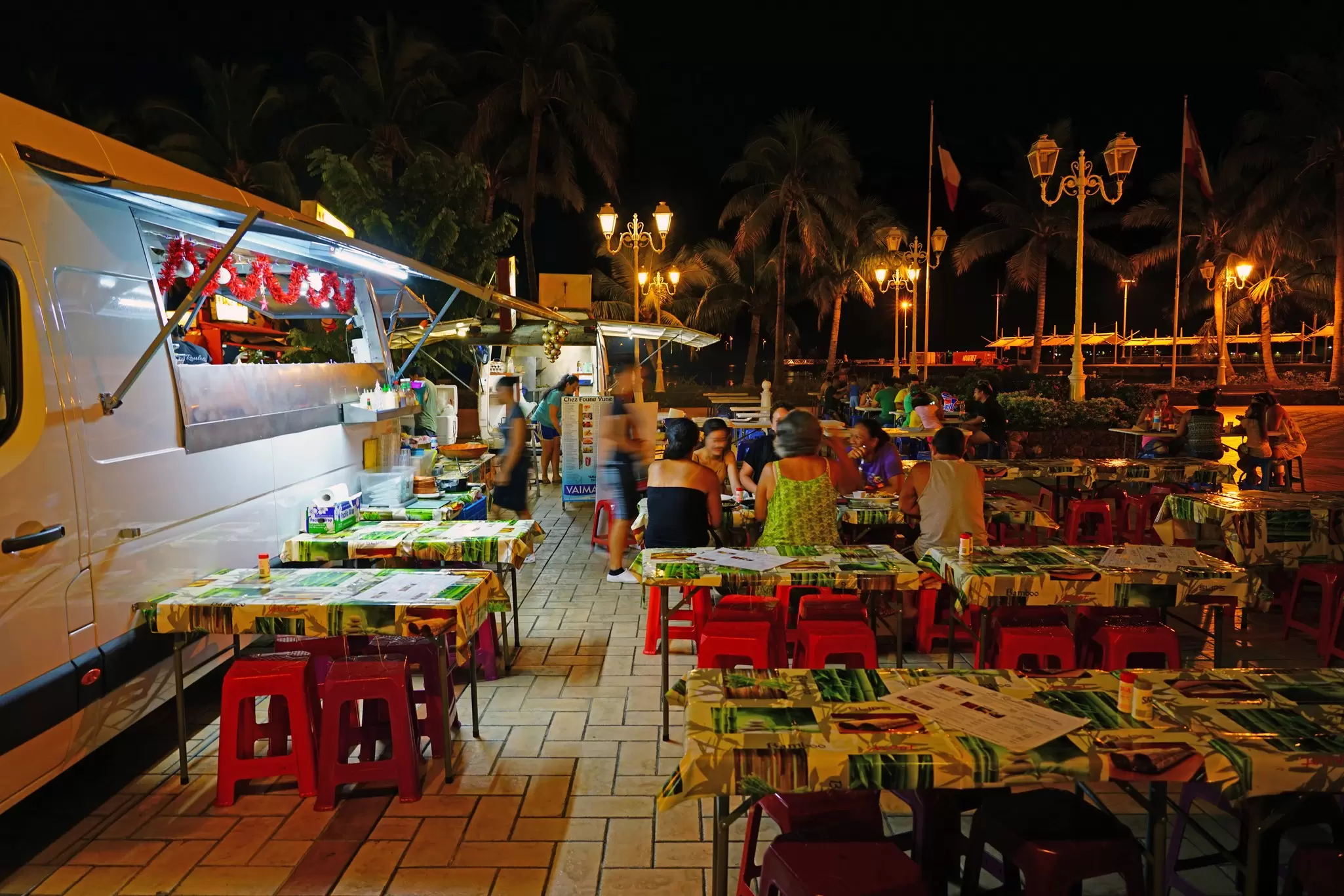 Tables covered with tablecloths and with bright plastic chairs are set up next to a food truck at night, Papeʻete, Tahiti