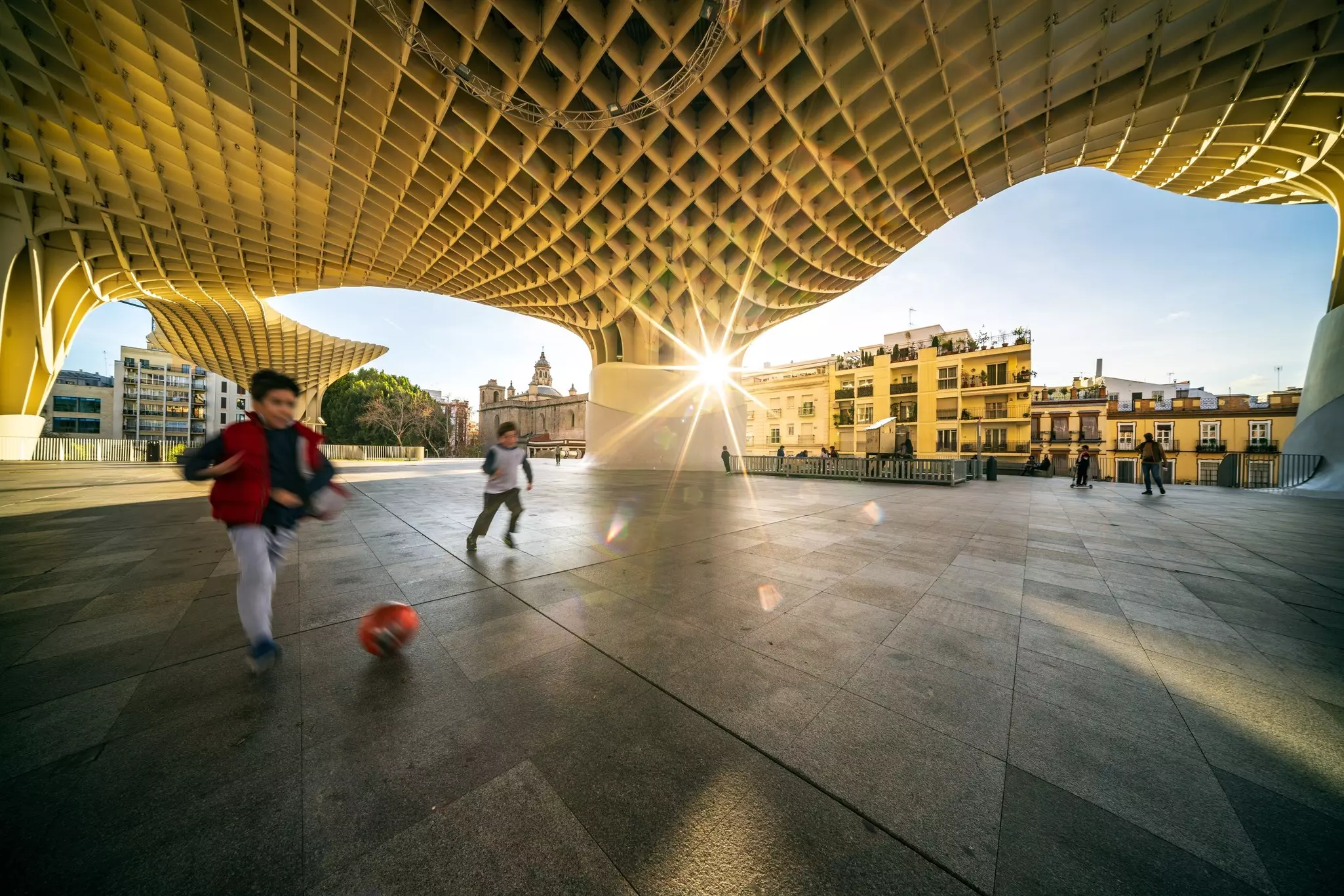 Children play in a plaza under a lattice structure as the rays of sunset light the scene.