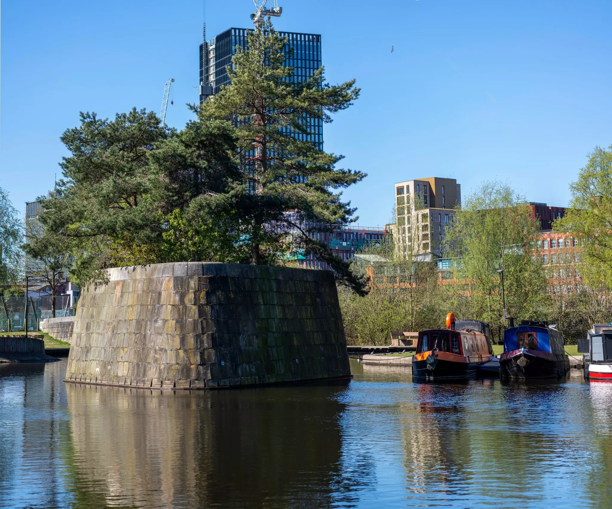 Image of canal with large cement structure in the middle holding pine trees, canal boats parked to the right of the image and city buildlings, includin a skyscraper, in the distance.