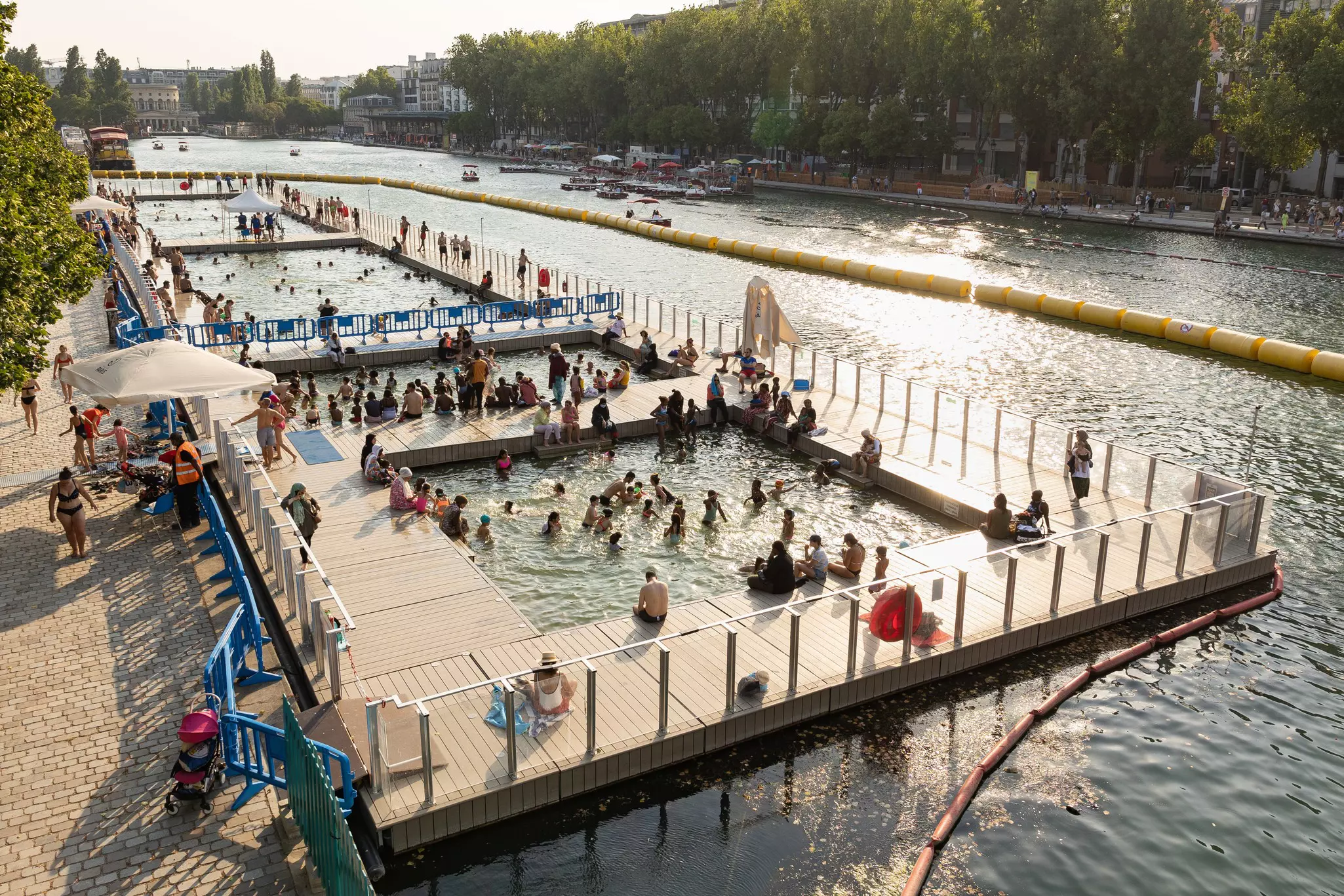 People swim and sit on docks on an artificial body of water adjacent to a wide river.