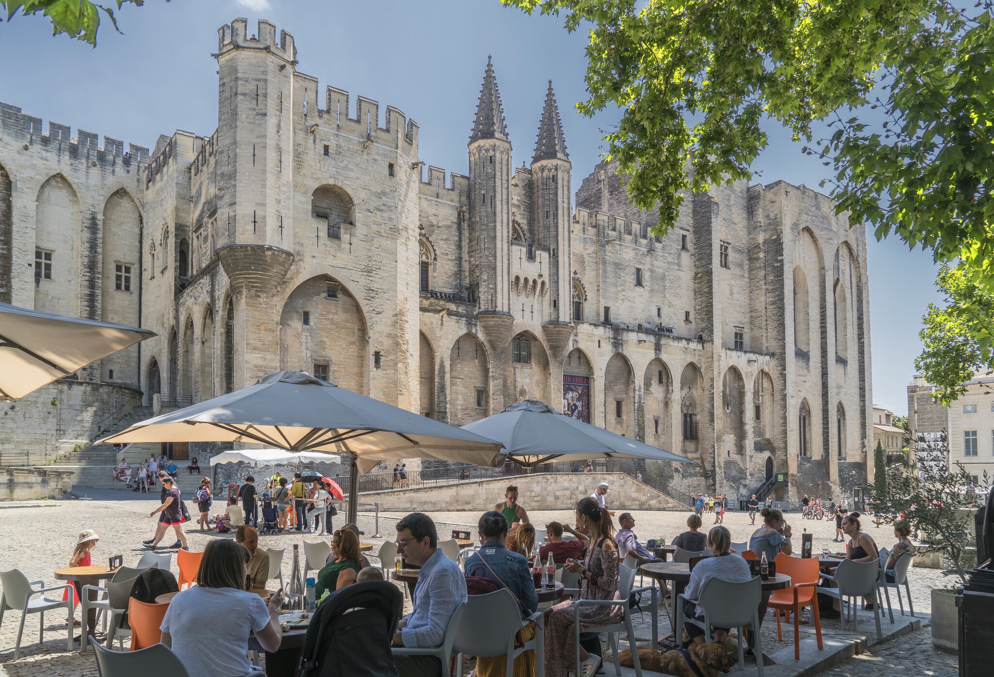 People sit in shade at an outdoor restaurant across from a Gothic structure in the sun.