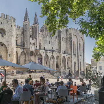 People sit in shade at an outdoor restaurant across from a Gothic structure in the sun.