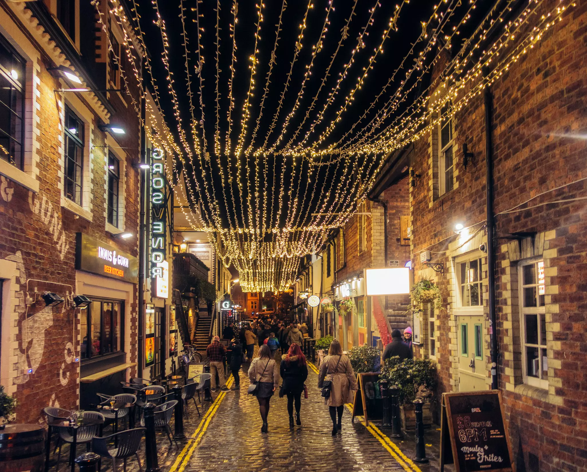 People walk through a wet cobblestone street lined with bars and restaurants; white lights are strung overhead.