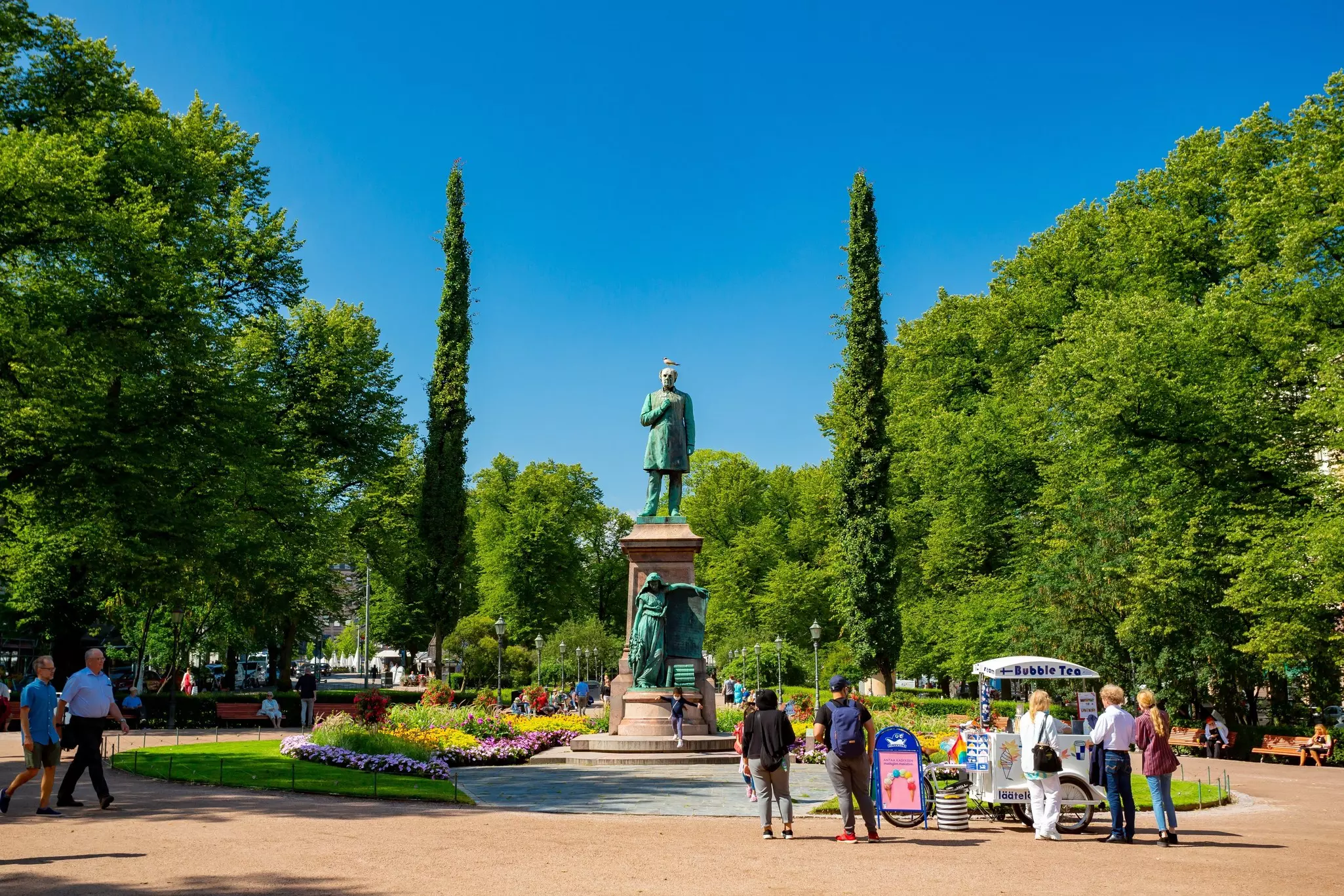 People wander through parkland near a large bronze statue of a man on a summer's day; Esplanadi Park, Helsinki
