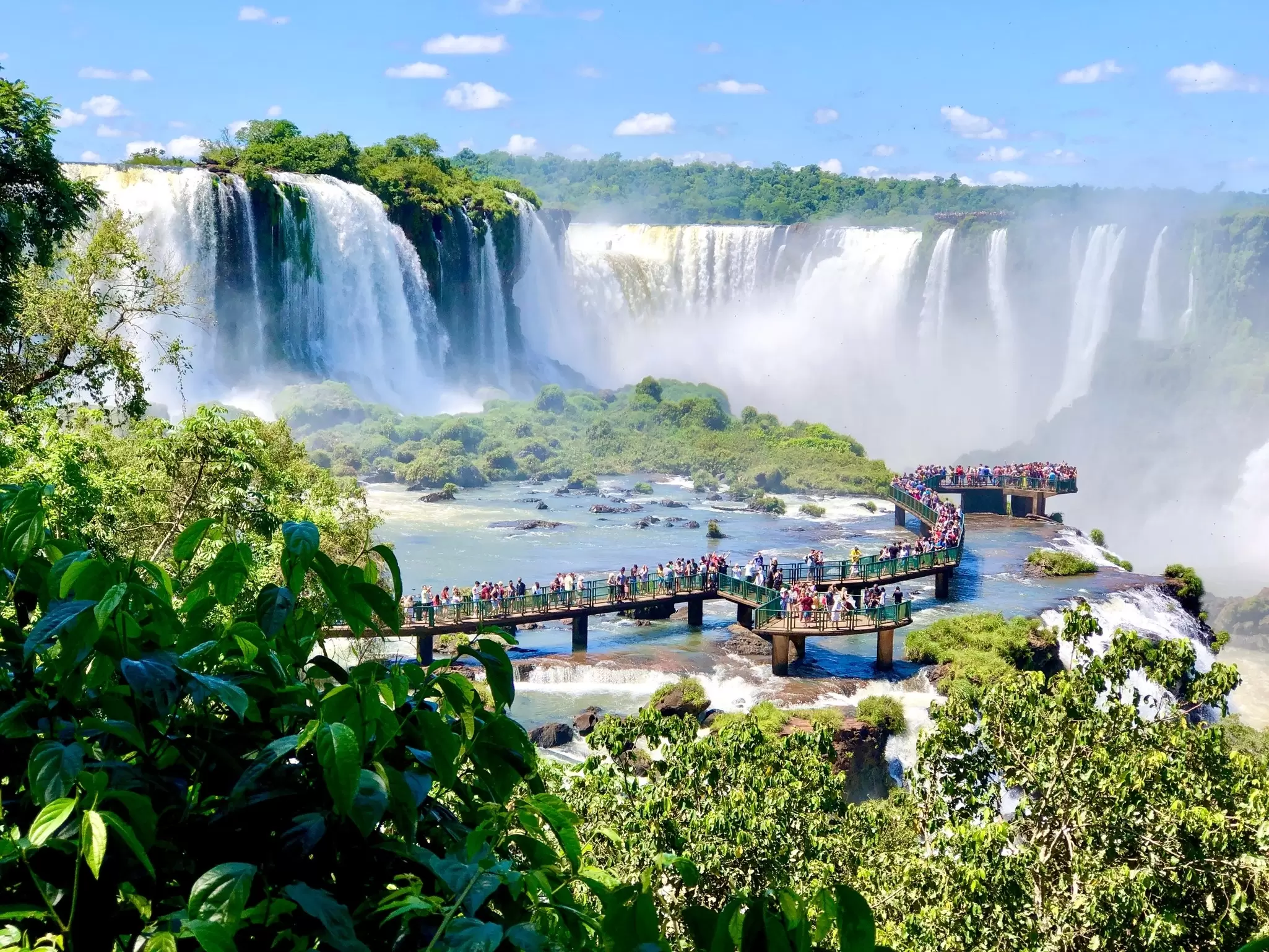 People on a boardwalk watching the cascades at Iguaçu Falls, Brazil, with spray rising from several large waterfalls.