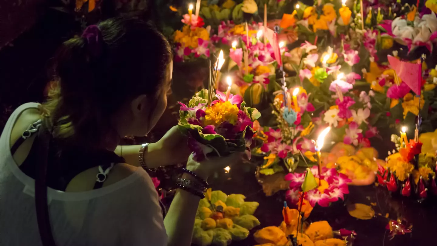 Woman puts flowers with candles stuck in them in a body of water with other similar flower and candle bundles.