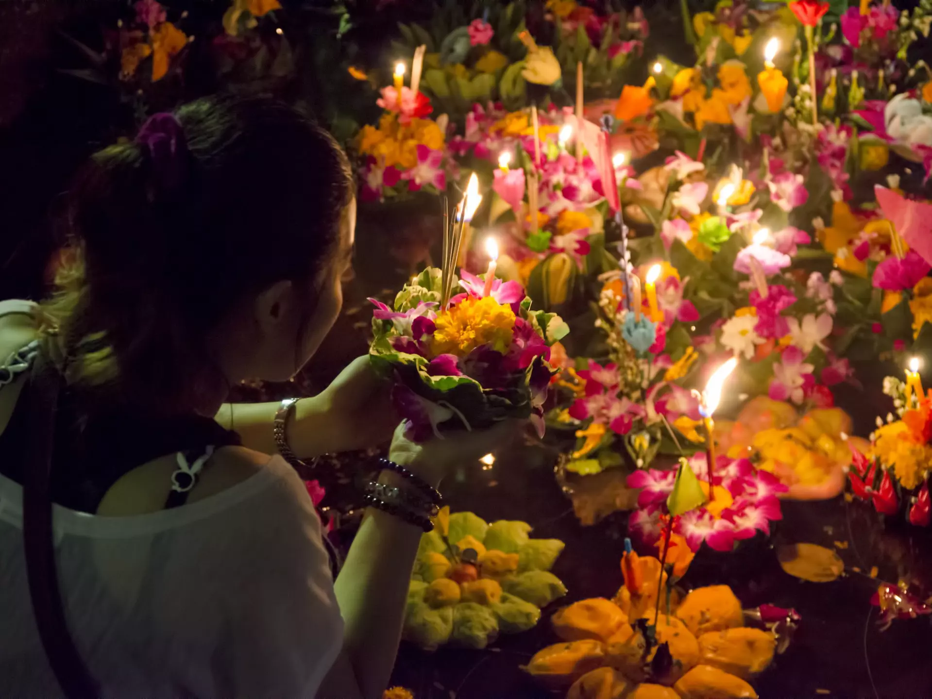 Woman puts flowers with candles stuck in them in a body of water with other similar flower and candle bundles.
