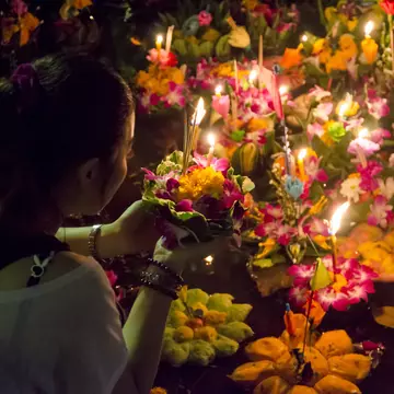 Woman puts flowers with candles stuck in them in a body of water with other similar flower and candle bundles.