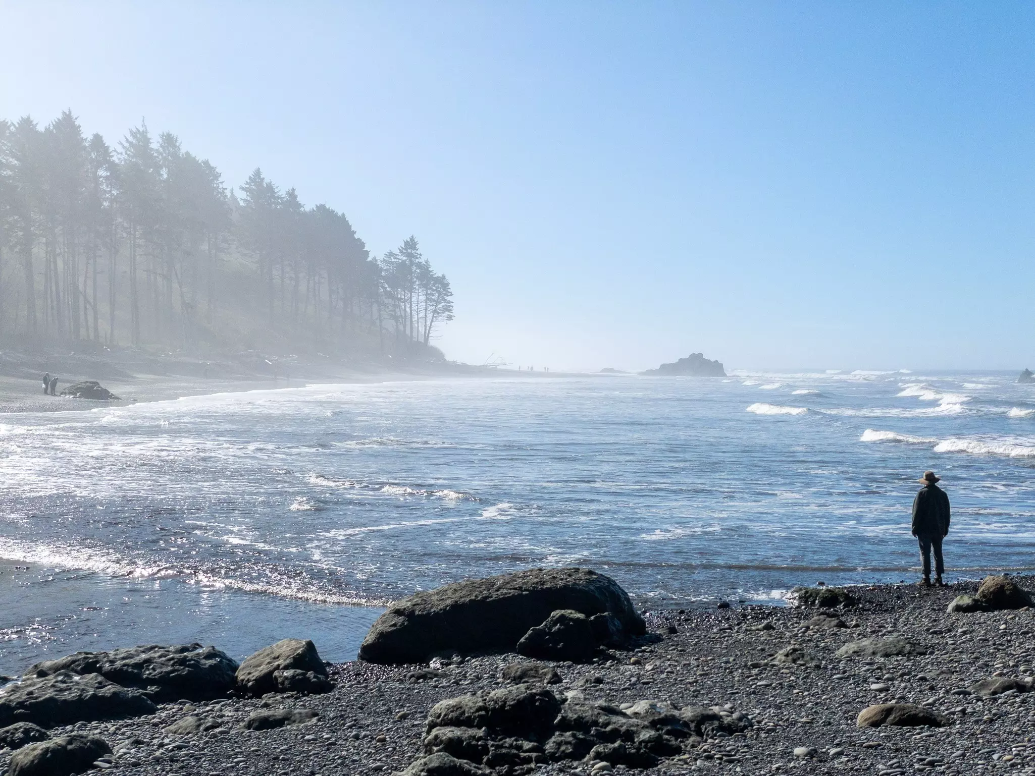 A person standing on a rocky shore looking at crashing surf.