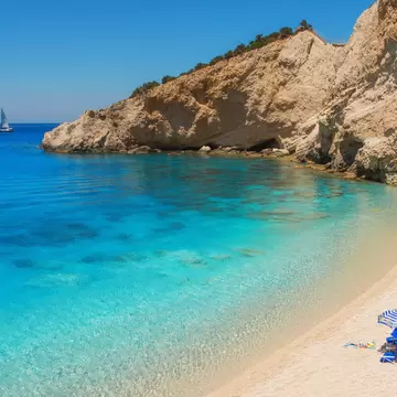 A white sand beach with a striped umbrella beside clear turquoise water, below pale limestone cliffs, a sailboat offshore.