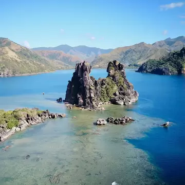 Aerial view of a dark limestone rock tower rising from a turquoise reef and lagoon, Hienghène, New Caledonia