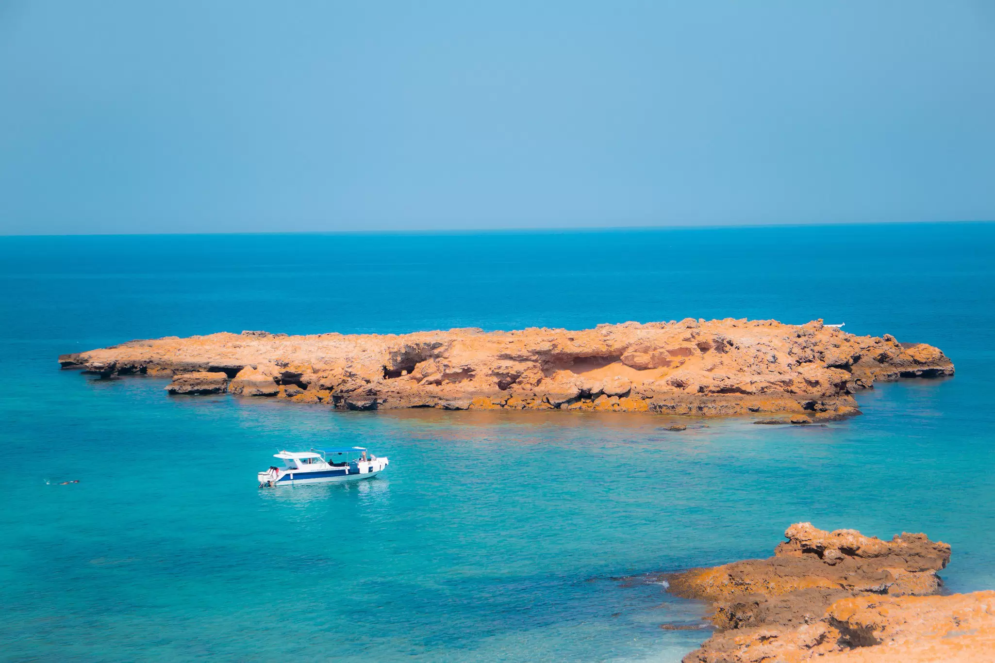 A small speed boat moored in turquoise ocean offshore of a rocky island; Damaniyat, Oman