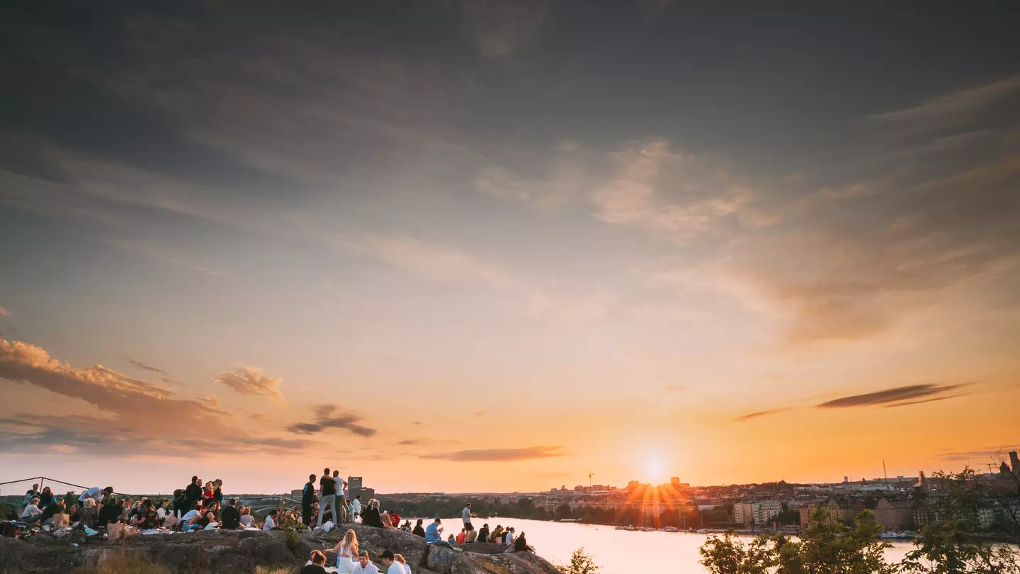 Groups of people sat on a cliff top looking out over a city at sunset
