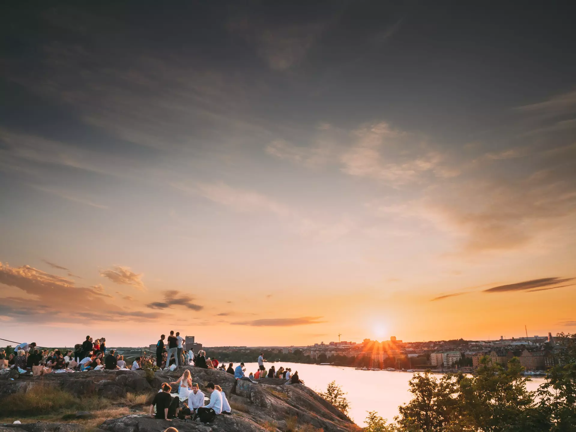 Groups of people sat on a cliff top looking out over a city at sunset
