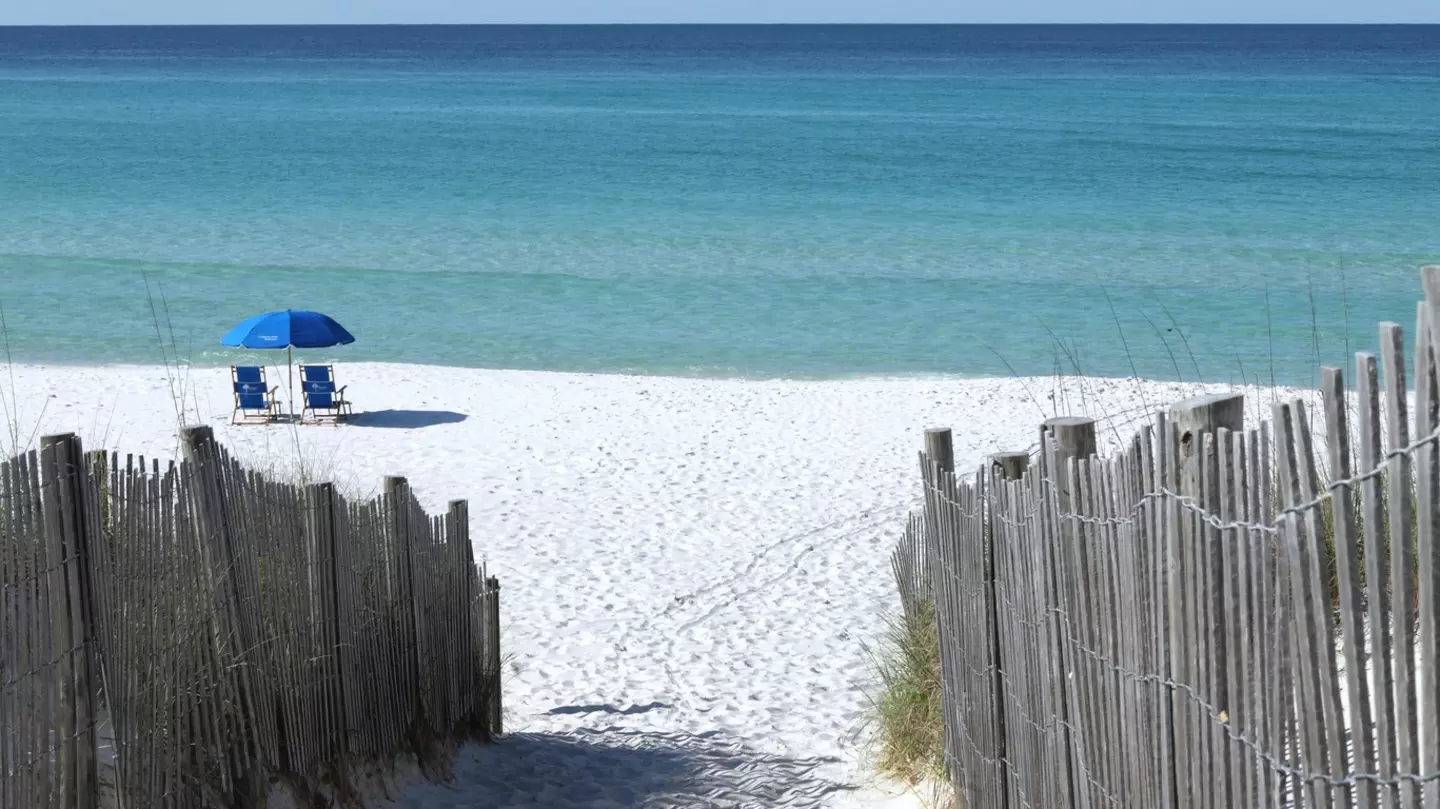 Wooden fences line the path to a white sand beach in Florida where two blue chairs sit under a blue umbrella.