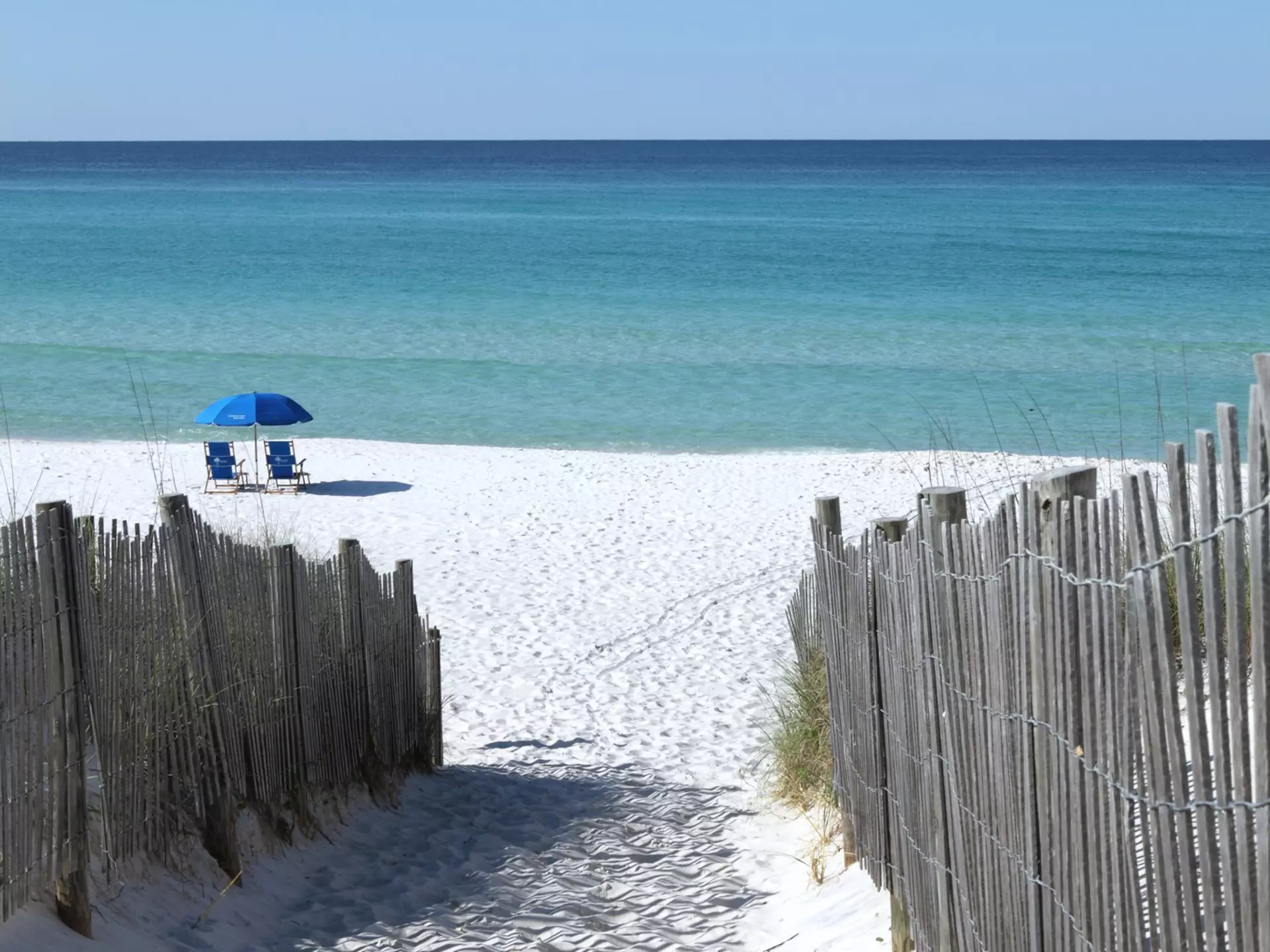 Wooden fences line the path to a white sand beach in Florida where two blue chairs sit under a blue umbrella.