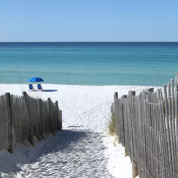 Wooden fences line the path to a white sand beach in Florida where two blue chairs sit under a blue umbrella.