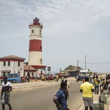 A red and white banded lighthouse standing over a busy coastal junction of minibuses, market stalls and people crossing the road.