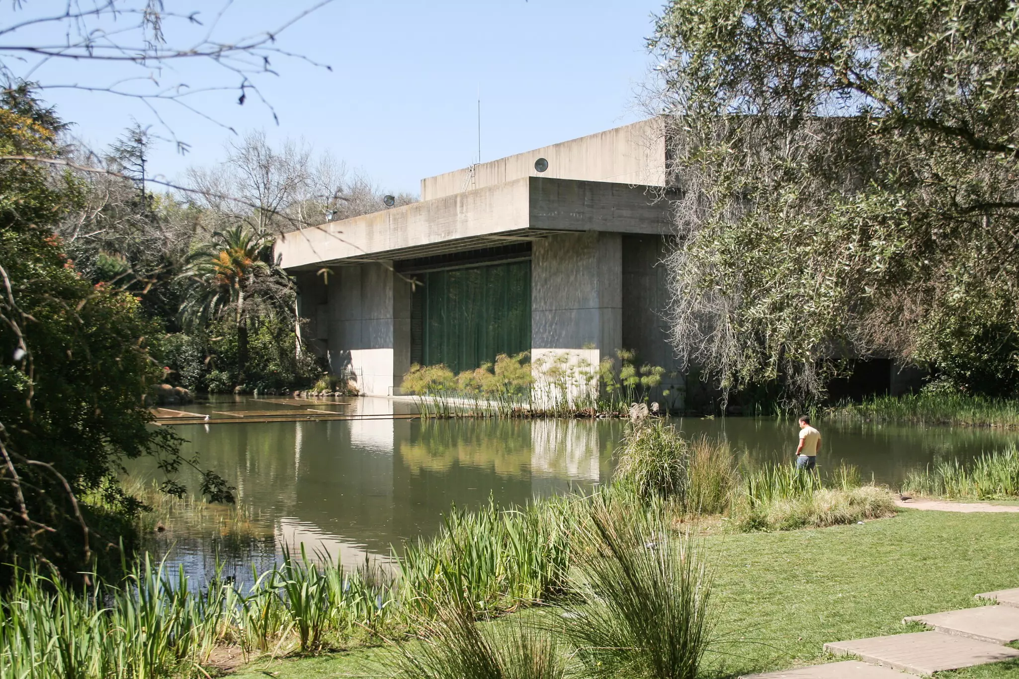 A brutalist-style concret building at the edge of a pond in a large garden.