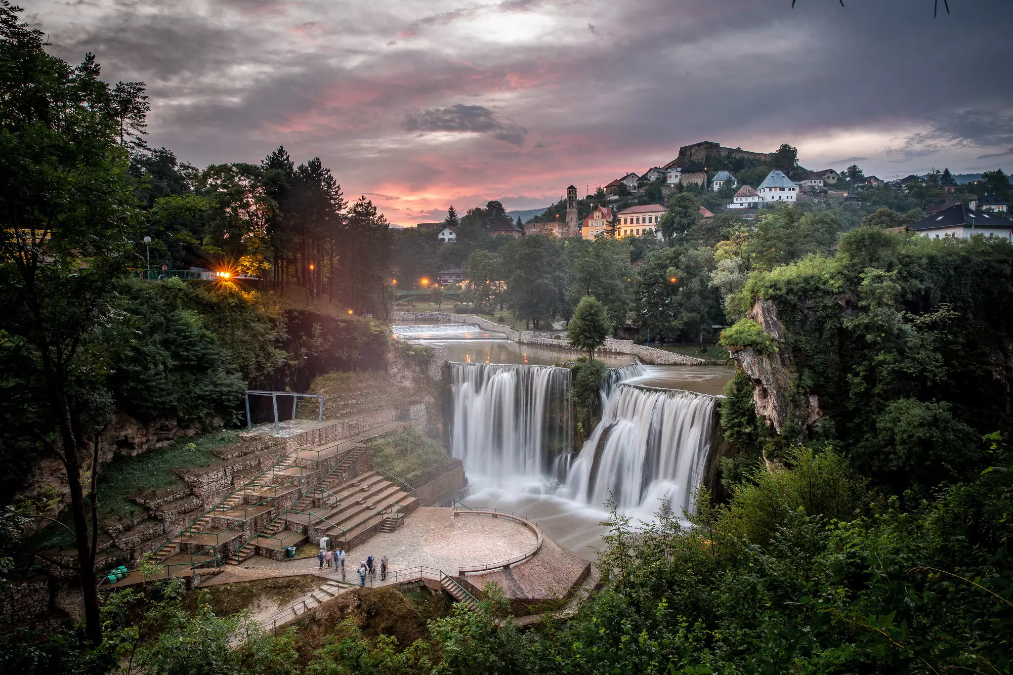 People gather on a platform for a view of a waterfall in a hilltop town at sunset.