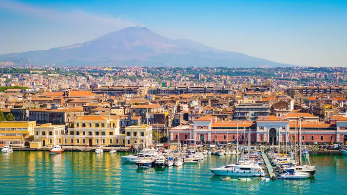 A view of a seaside town and harbor in Italy; the low buildings are in pastel shades, and there is a large mountain in the background.