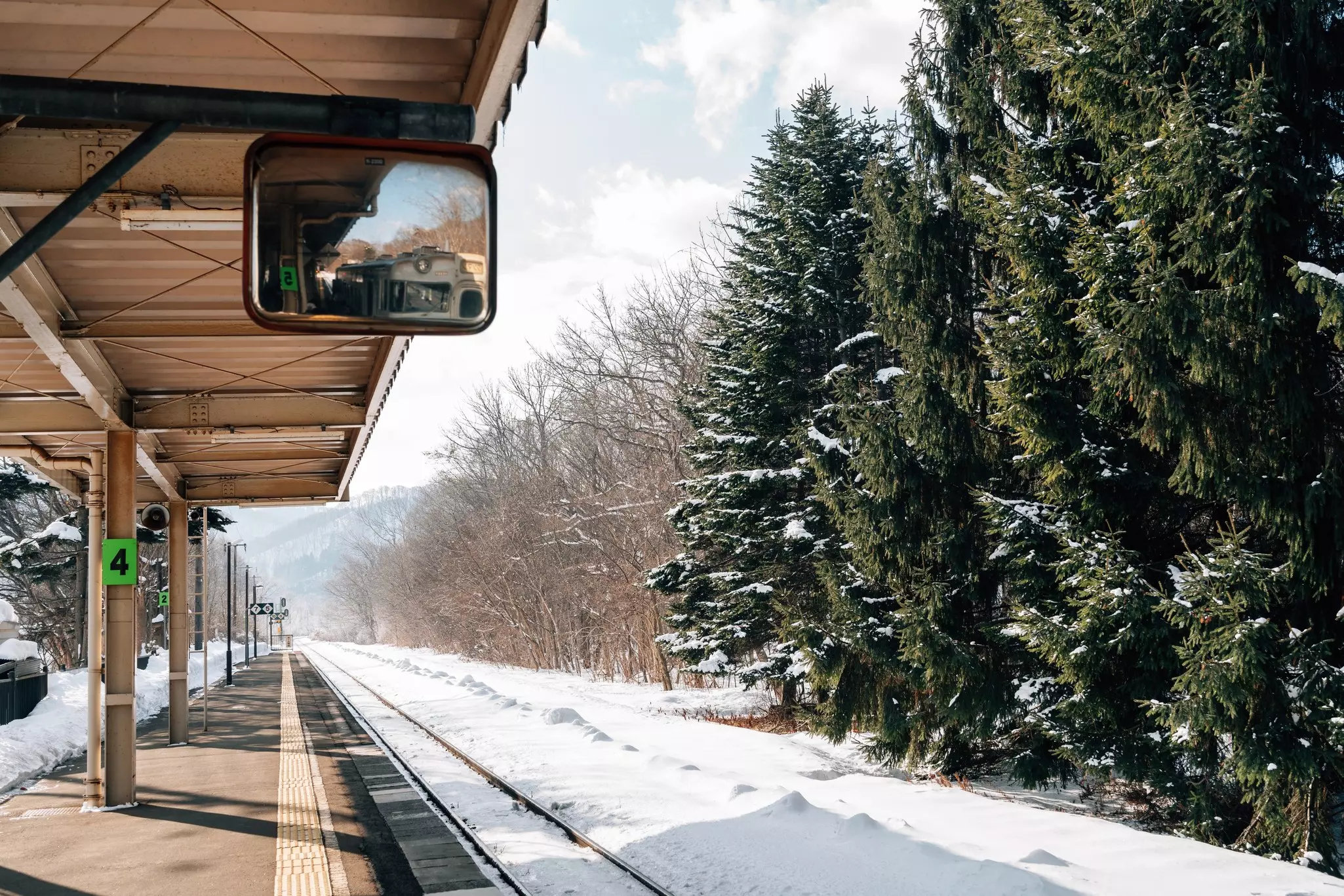 Snow covers a railway by a station platform. Snow-covered pines and other trees lie across the tracks, and a train can be seen reflected in a small mirror hanging above.
