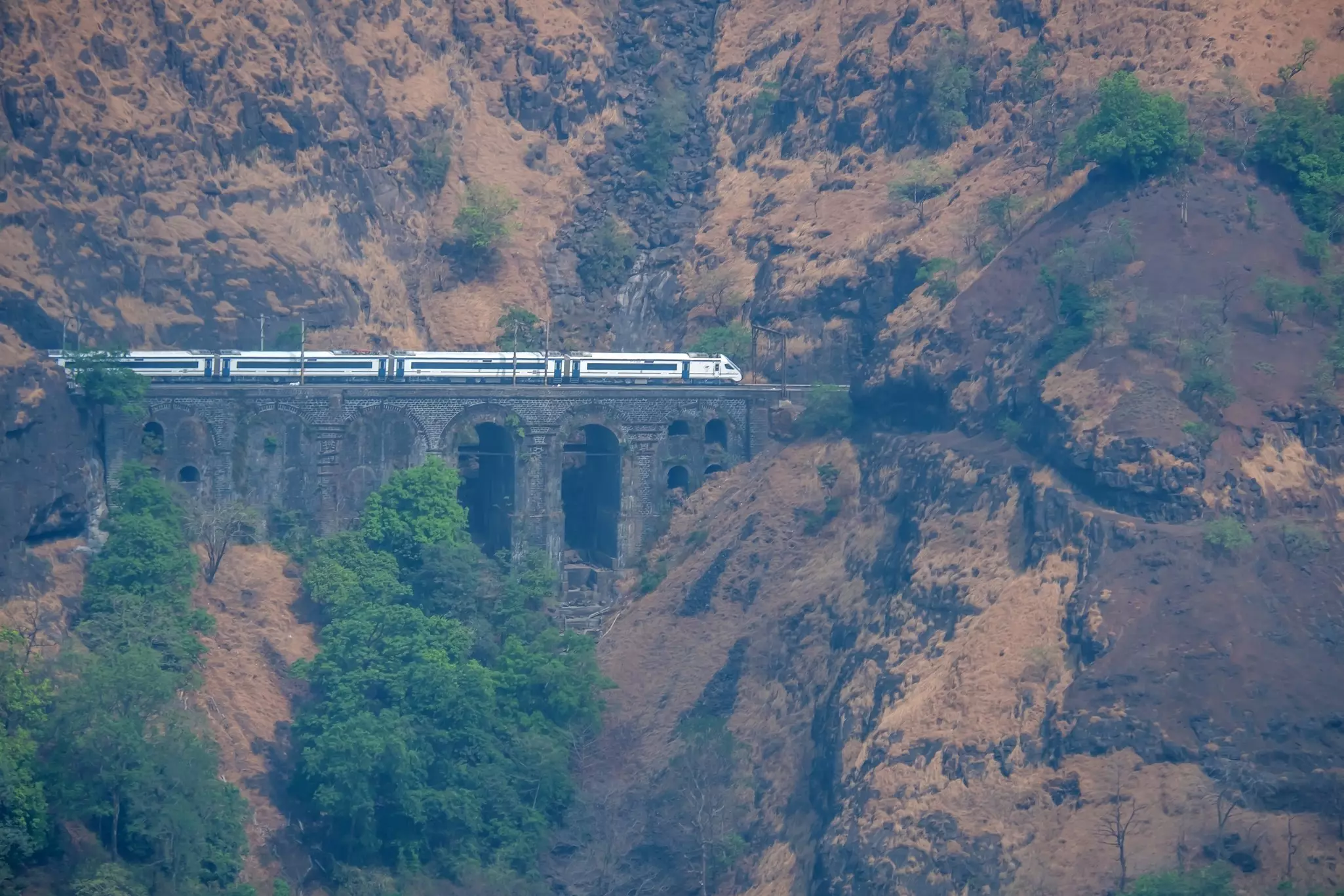 A train with white cars travels on a stone viaduct along a mountainside.