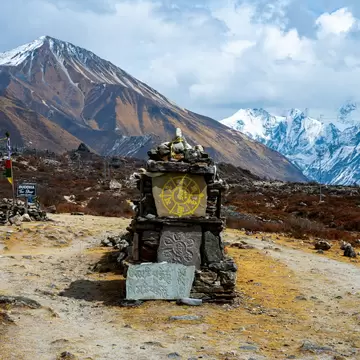 Stone shrine carved with Buddhist symbols on a dirt trail, brown hills and snowy Himalayan peaks behind, Langtang Valley