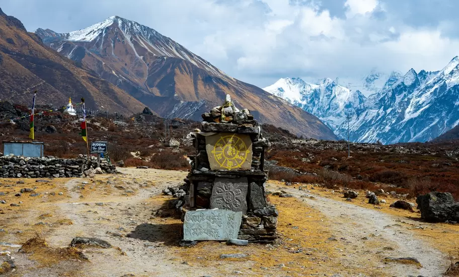 Stone shrine carved with Buddhist symbols on a dirt trail, brown hills and snowy Himalayan peaks behind, Langtang Valley