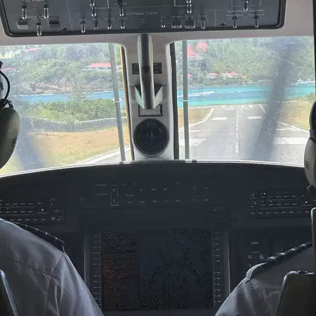 View over the shoulder of the pilots looking through cockpit. The short runway ahead of them ends in blue ocean waters.