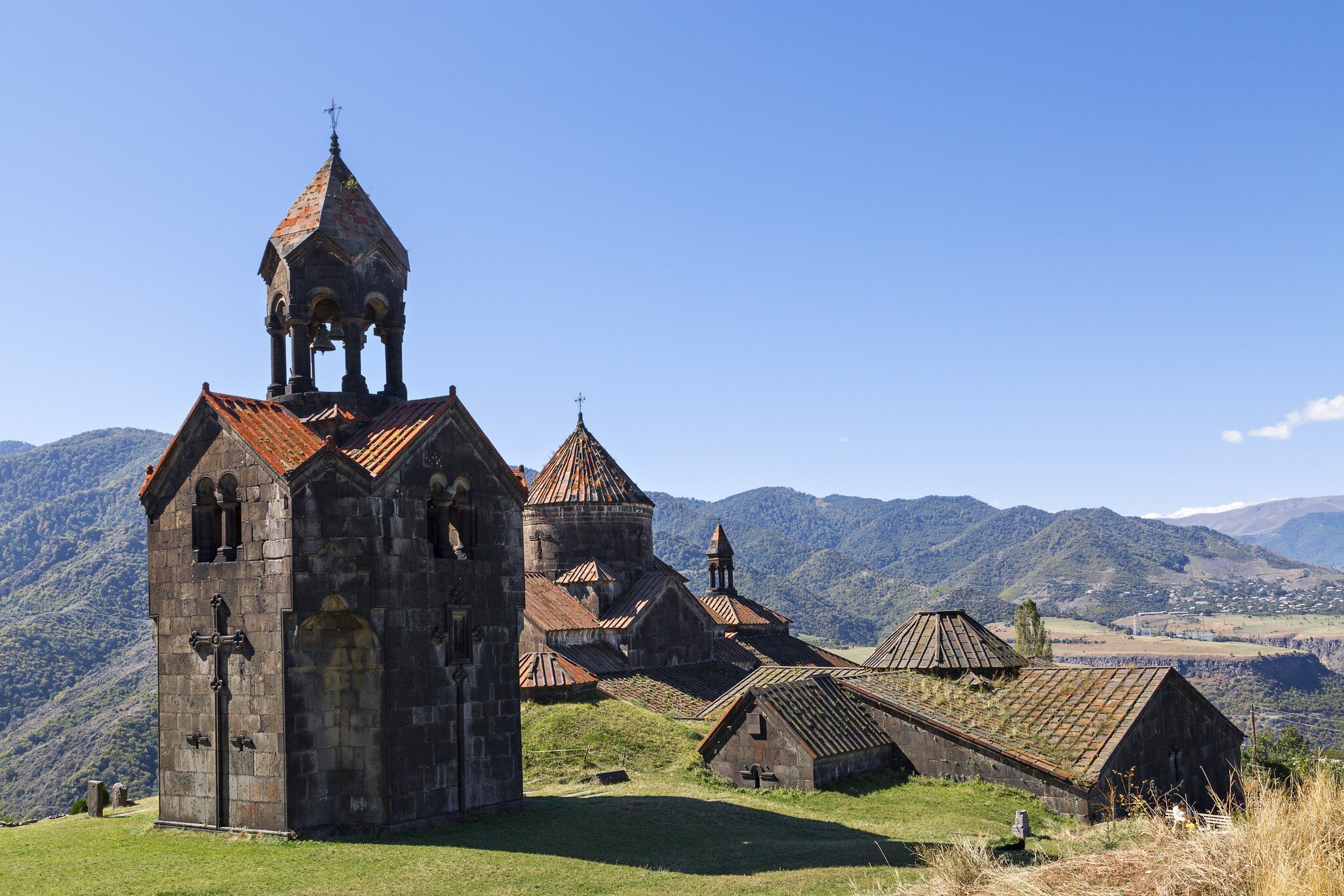 A dark stone bell tower with red-tiled roofs stands before domed church buildings and forested mountains.