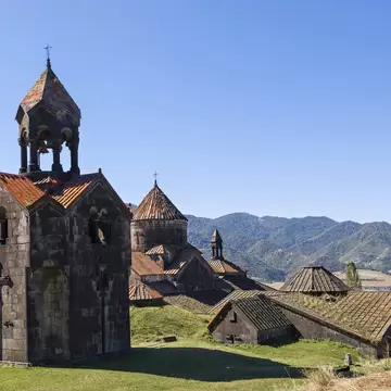 A dark stone bell tower with red-tiled roofs stands before domed church buildings and forested mountains.