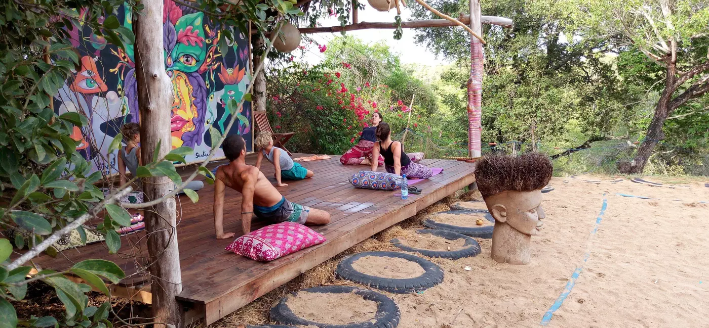 A small group of people follow a yoga teacher in a class taught in an outdoor shelter.