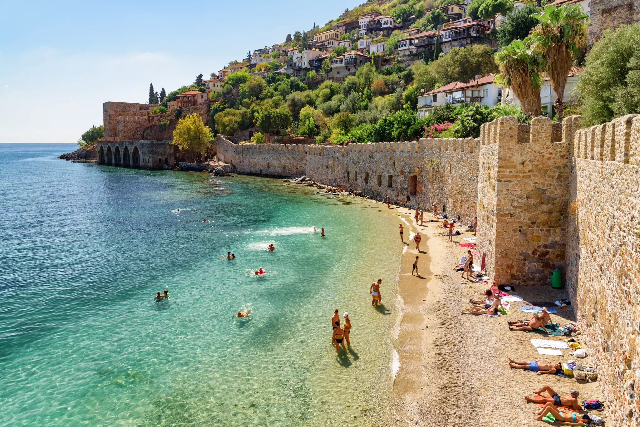 View clear blue ocean with people swimming, sunbathers on a narrow strip of beach near a high stone wall, and houses above on a sunny day.