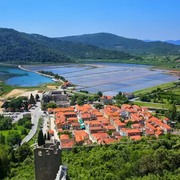Red-roofed houses beside rectangular salt pans and a bay, with green hills behind and a stone wall tower, in Ston, Croatia