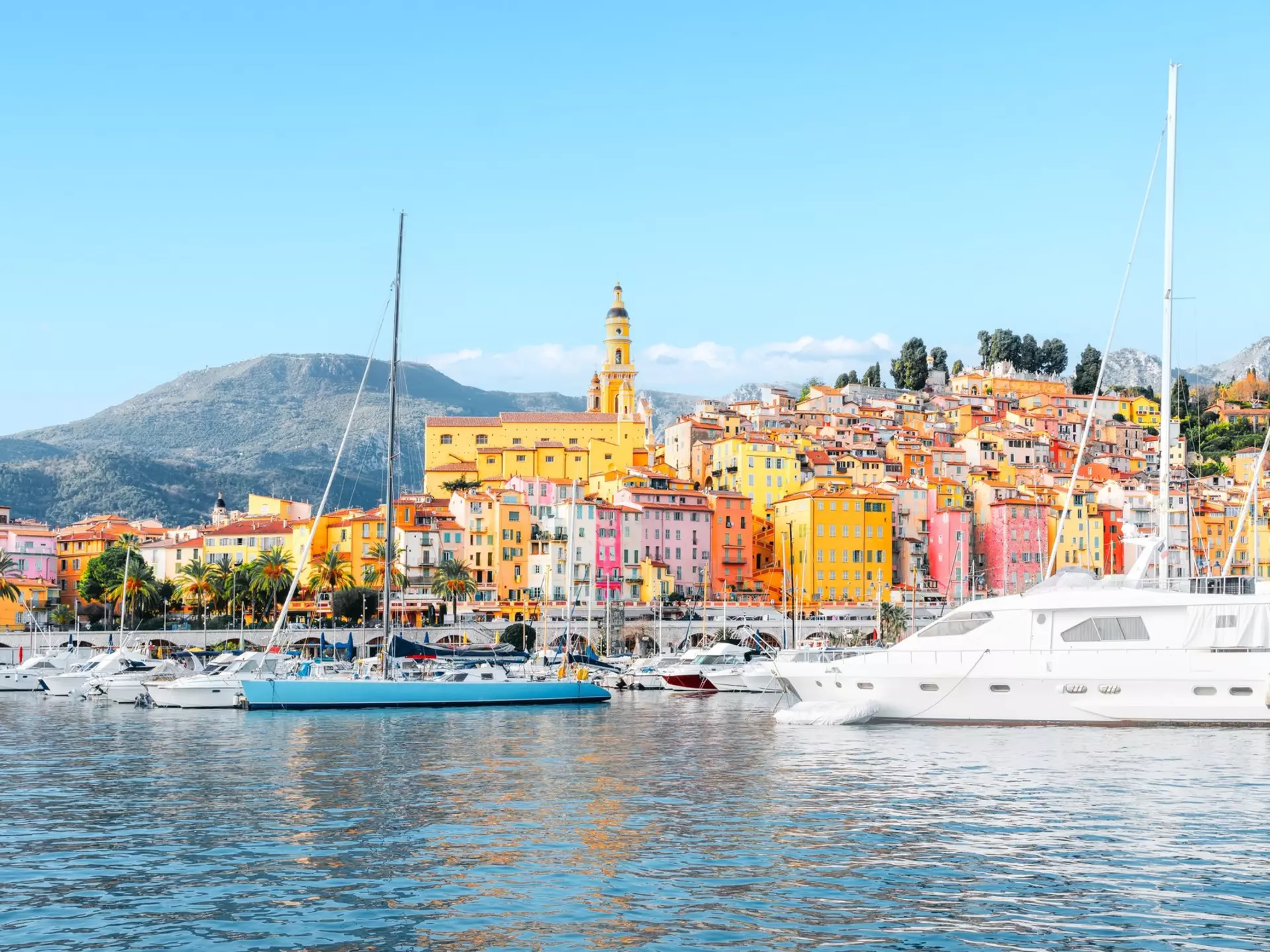 Pastel-colored buildings bathed in sunlight stacked on the edge of a hill near a small harbor full of sailing boats and expensive yachts.