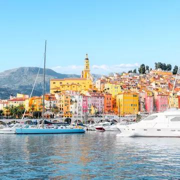 Pastel-colored buildings bathed in sunlight stacked on the edge of a hill near a small harbor full of sailing boats and expensive yachts.