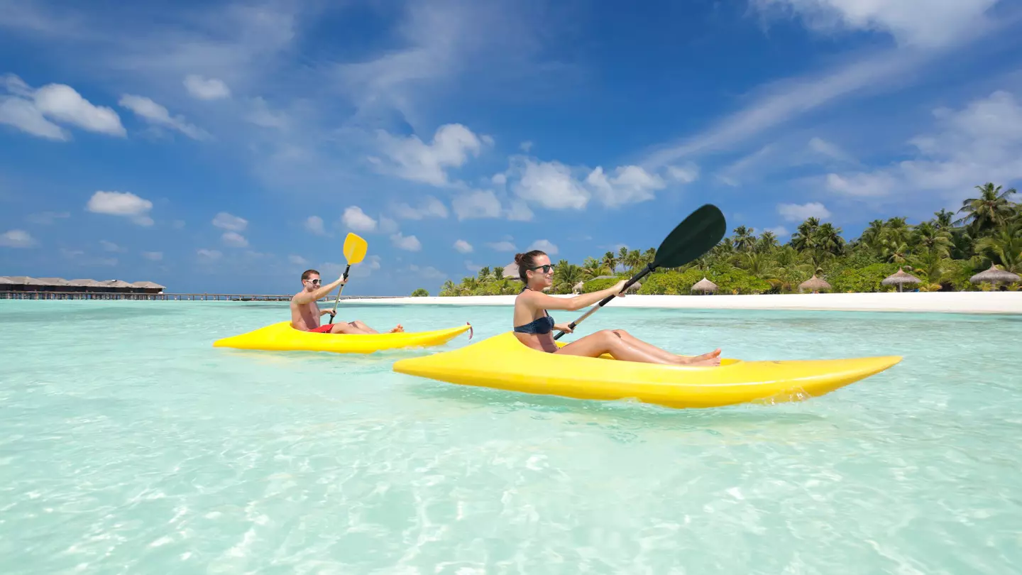 A couple in yellow kayaks paddle through crystal-clear water in the Maldives, with blue skies above them and a row of sunshades in the white sand in the distance.