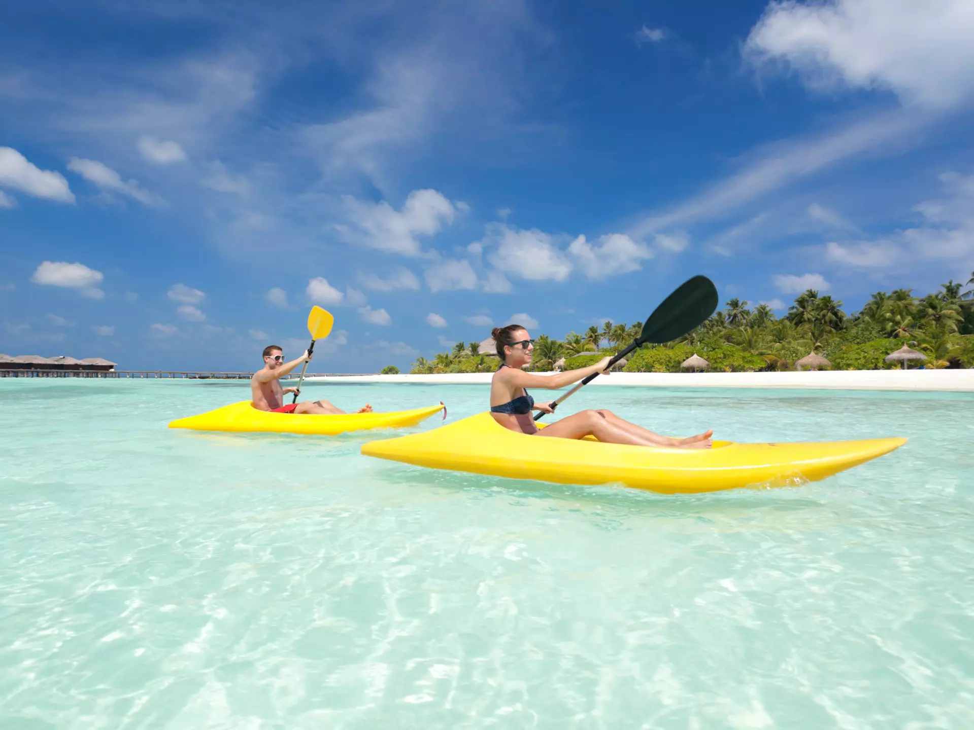 A couple in yellow kayaks paddle through crystal-clear water in the Maldives, with blue skies above them and a row of sunshades in the white sand in the distance.