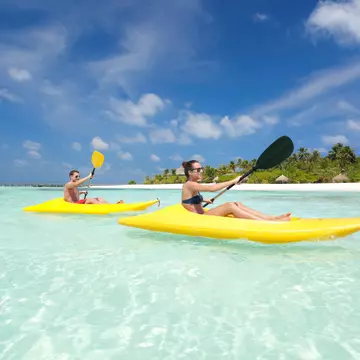 A couple in yellow kayaks paddle through crystal-clear water in the Maldives, with blue skies above them and a row of sunshades in the white sand in the distance.